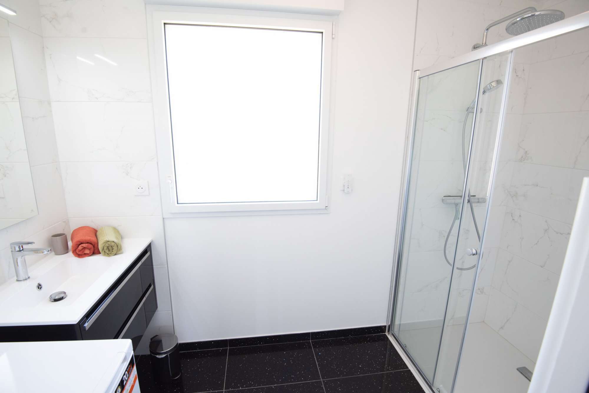 A modern bathroom with a glass shower enclosure, a white sink, and a window above the mirror.