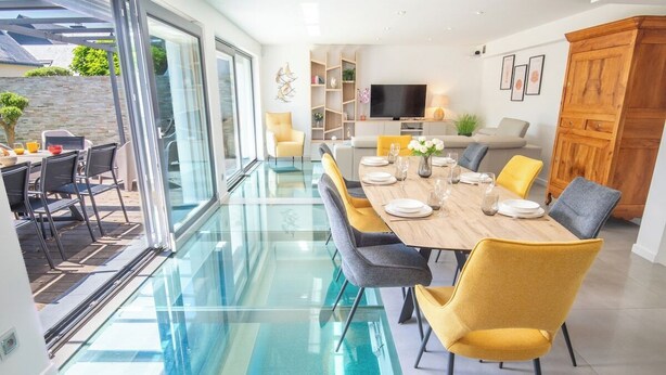 A modern dining area with a glass floor leading to an outdoor patio, a dining table set with chairs, and a television in the background. - Luxury villa with indoor pool, 5 minutes from the sea via Groom* (Larmor-Plage)