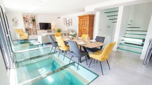 A modern dining area with a glass floor leading to a pool, surrounded by yellow and gray chairs, a wooden table, and a TV on the wall. - Luxury villa with indoor pool, 5 minutes from the sea via Groom* (Larmor-Plage)