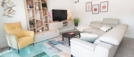 A modern living room with a glass floor, white sofa, yellow armchair, wooden shelving unit, flat-screen TV, and decorative wall art.