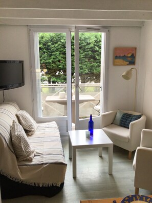 A living room with a sofa, a smaller armchair, a coffee table, and a TV. There is a large window with a view of greenery outside. - Cozy Apartment on the Corniche Vendéenne   (Saint-Hilaire-de-Riez)
