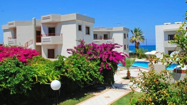 A resort with multiple white buildings, a swimming pool, and lush greenery. - John Mary Apartments by Estia (Hersonissos)