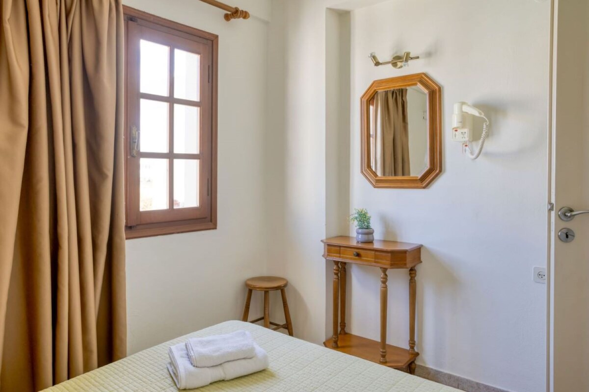 Une chambre à coucher avec un lit, une table de chevet en bois, un miroir et une fenêtre avec des rideaux.