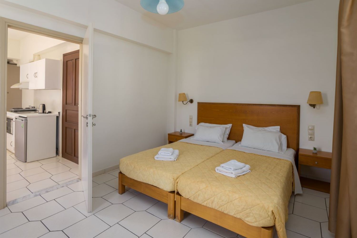 A hotel room with a double bed, two bedside tables, a small kitchenette, and a bathroom visible through an open door.