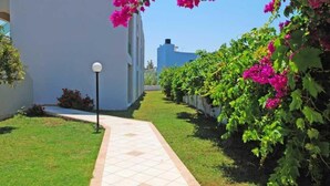 A pathway leading to a building with a tiled entrance, surrounded by lush greenery and vibrant pink flowers. - John Mary Apartments by Estia (Hersonissos)