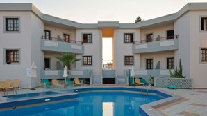 A pool area with lounge chairs and umbrellas, surrounded by buildings with balconies. - John Mary Apartments by Estia (Hersonissos)