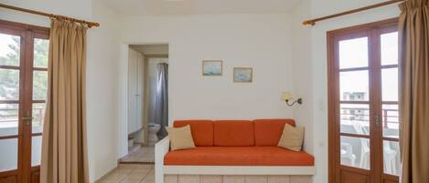 Une pièce avec un sol carrelé de couleur beige, un canapé blanc agrémenté de coussins orange, deux tableaux encadrés accrochés au mur et une fenêtre avec des rideaux.