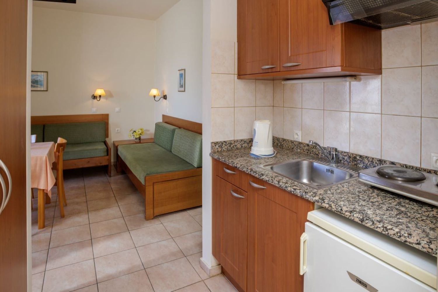 A compact kitchen with a sink, stove, and wooden cabinets. There is a dining area with a table and chairs, and a small sofa.