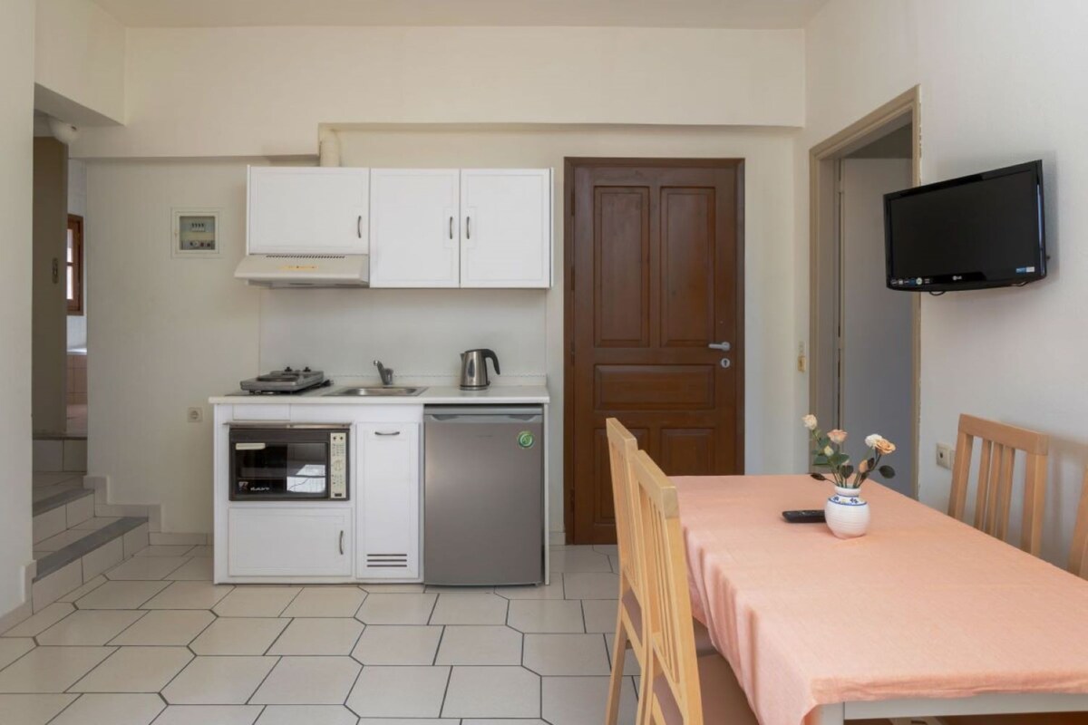 Eine kleine Küche mit weißen Schränken, einem eingebauten Ofen und einem kleinen Kühlschrank. Im Vordergrund steht ein Holztisch mit einer rosa Tischdecke und einem Blumenstrauß.