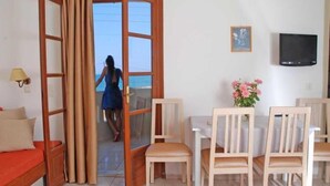 A room with a bed, a dining table with chairs, a TV on the wall, and a view of the sea through an open door. - John Mary Apartments by Estia (Hersonissos)