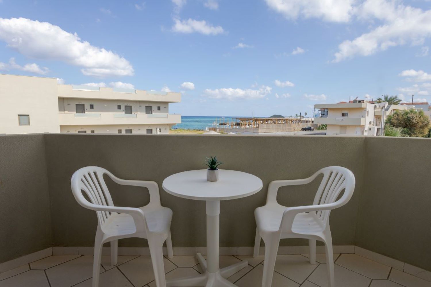 Un balcon avec deux chaises en plastique blanches et une petite table, donnant sur une plage et des immeubles.