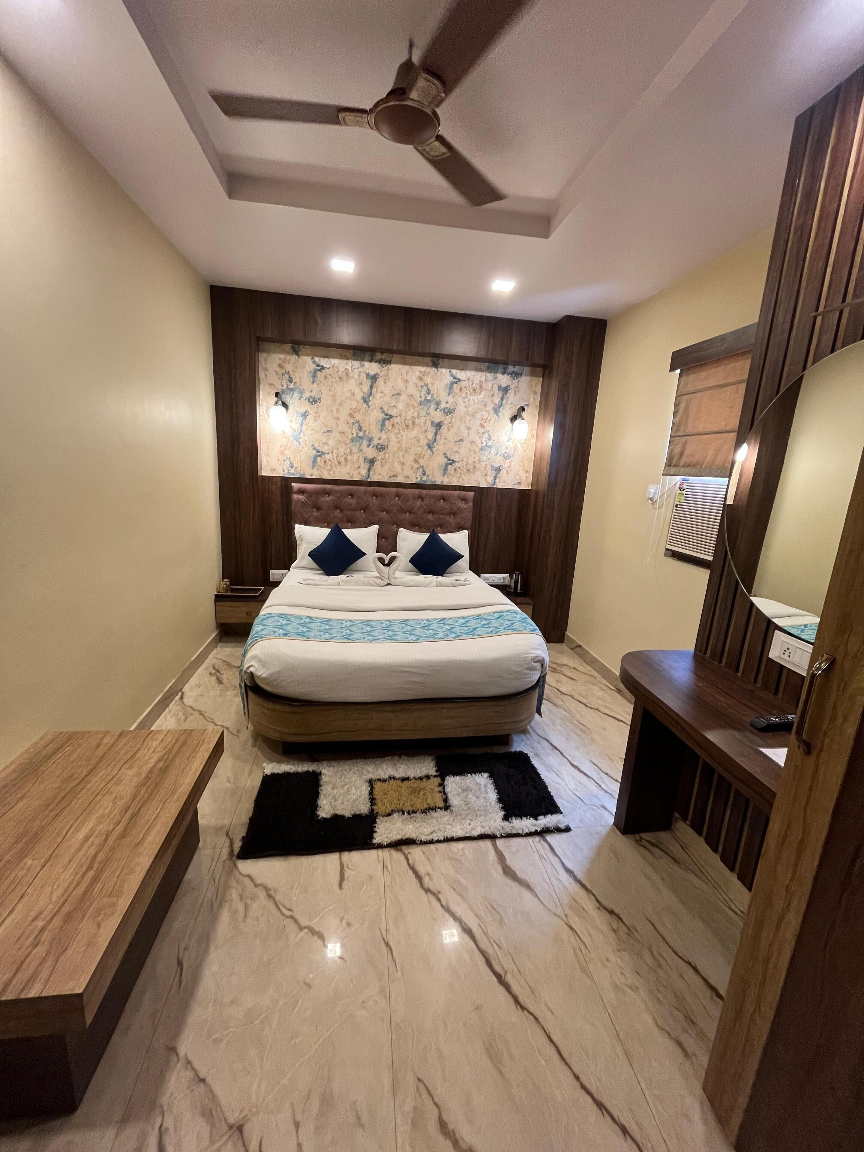 A bedroom with a bed, a ceiling fan, a nightstand, a bedside table, a wooden bench, a marble floor, and a wall-mounted lamp.