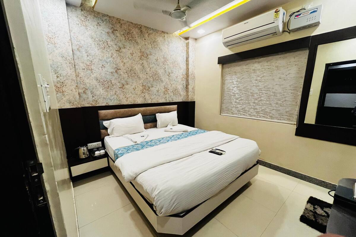 A hotel room with a bed, a nightstand, an air conditioning unit, and a window with a view.