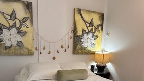 A neatly made bed with a white quilt and a green throw pillow, flanked by two wall-mounted paintings of flowers. - First Floor Unit with big patio. Countryside steps away from beautiful beaches. (Cabo Rojo)