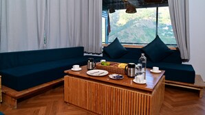 A room with a sofa, a wooden coffee table with breakfast items, and a view of mountains through a window. - Valtara Resort (Nainital)