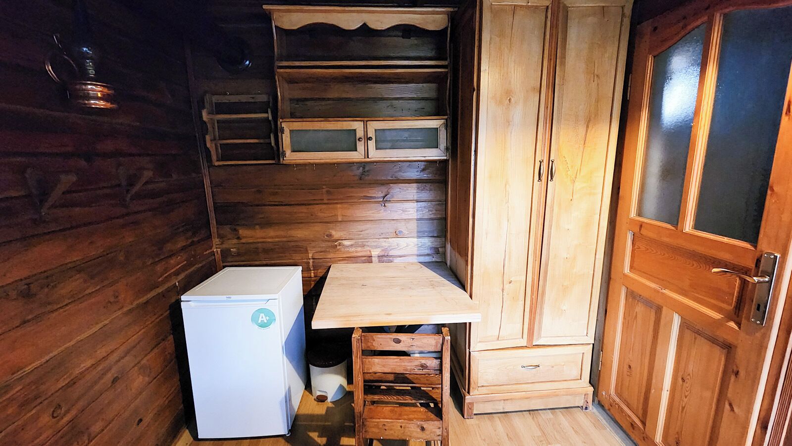 A compact kitchen with wooden walls, a white refrigerator, a wooden table, and a cabinet with a glass door.