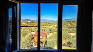 Vista a través de una ventana de una cabaña de madera con techo rojo, rodeada de vegetación densa y montañas al fondo.
