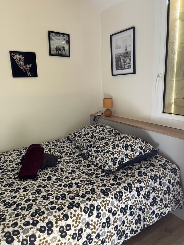 Un dormitorio con una cama, dos cuadros en la pared, una mesita de noche con una lámpara y una ventana con vistas.