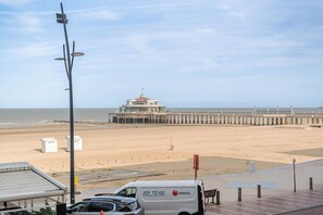 Une plage avec une longue jetée qui s’avance dans la mer, un fourgon blanc garé sur la promenade, et un bâtiment coiffé d’un dôme en arrière-plan.