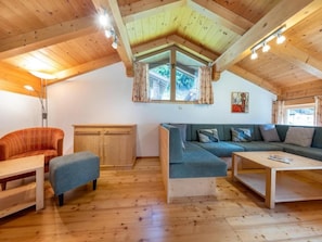 A cozy attic room with a wooden ceiling, a sofa, a chair, a small table, and a window with curtains.