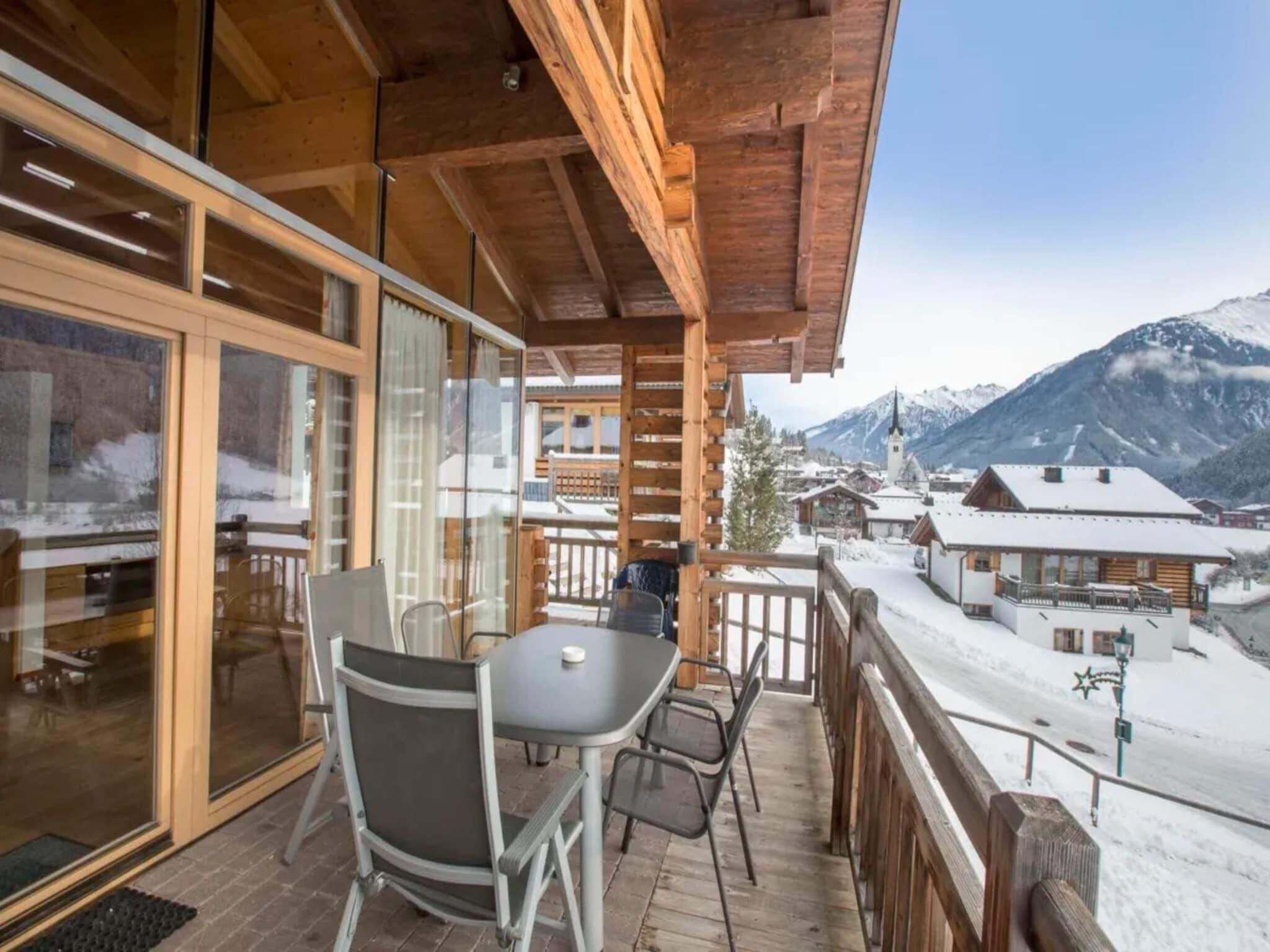 Ein Holzbalkon mit Tisch und Stühlen, mit Blick auf ein verschneites Dorf und Berge.