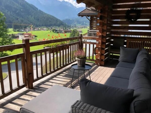 A balcony with a sofa and a small table, overlooking a green landscape and a mountain range.