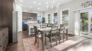 A modern kitchen with a dining area, featuring a chandelier, wooden cabinets, and a large window.