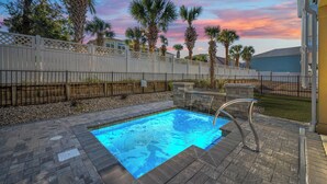 A swimming pool with a waterfall feature, surrounded by palm trees and a white fence.