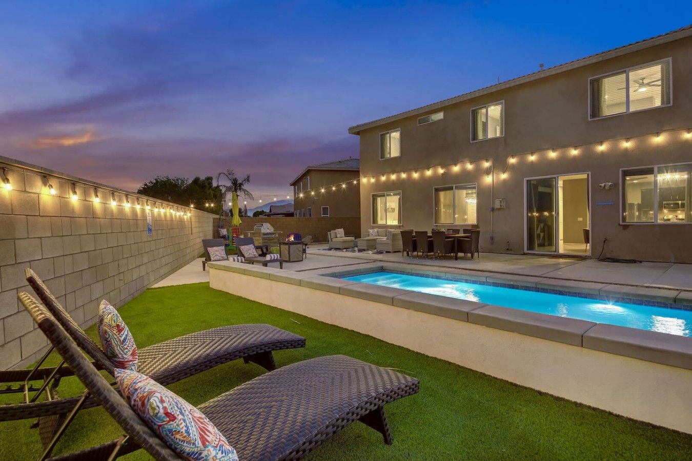 Un patio trasero con piscina, mobiliario exterior e iluminacion con guirnaldas.