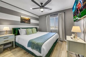 A hotel room with a bed, a ceiling fan, a painting, a window with curtains, and a nightstand with a lamp.