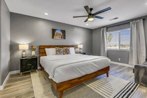 A bedroom with a bed, nightstands, a ceiling fan, a window with curtains, and a painting on the wall. - Wonders Nest: Pool, Spa, Arcades, Putting Green! (Coachella)