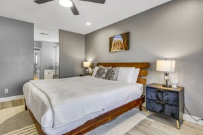 A bedroom with a large bed, a nightstand with a lamp, a ceiling fan, and a painting on the wall.