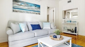 A living room with a grey sofa, a white coffee table, and a painting of a beach scene. - Charming Folly Beach Home - Perfect Location (Folly Beach)