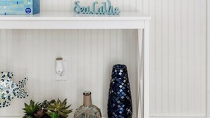A white interior with a mirror, a shelf with a 'Sea Calming' sign, a patterned planter, and a decorative vase. - Charming Folly Beach Home - Perfect Location (Folly Beach)