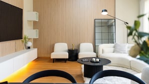 A modern living room with a wooden panel wall, a flat-screen TV, two white armchairs, a black coffee table, and a white sofa. - YouSleep Hostel (Dubai)