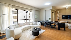 A modern living room with a sectional sofa, a round coffee table, a dining table with chairs, and a flat-screen TV mounted on the wall. - YouSleep Hostel (Dubai)