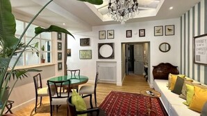 A well-lit living room with a chandelier, a dining area with a round table and chairs, a sofa with cushions, and a large plant. - Testaccio GreenHouse | ArtDesignApartments (Rome)