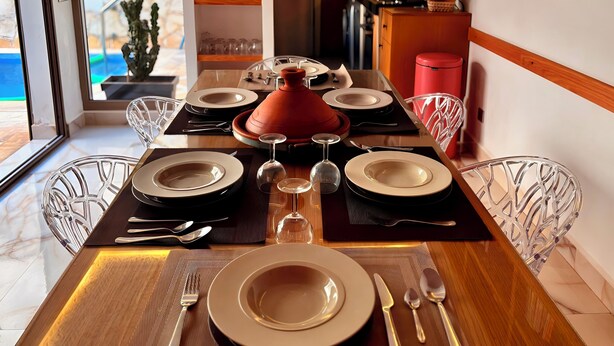 Une table à manger est dressée avec des assiettes, des couverts et un pot rouge. On aperçoit une piscine et une cuisine en arrière-plan.