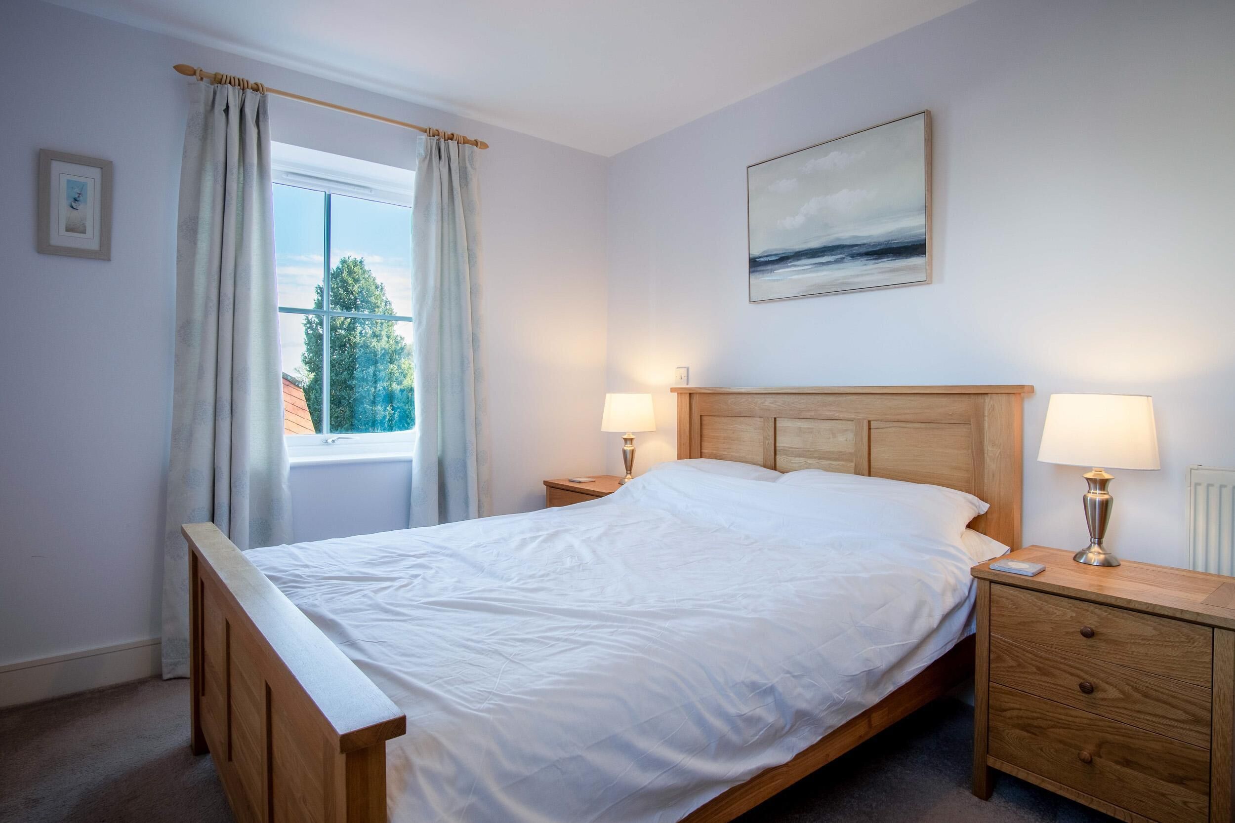 A bedroom with a wooden bed, two bedside tables with lamps, a window with curtains, and a painting on the wall.