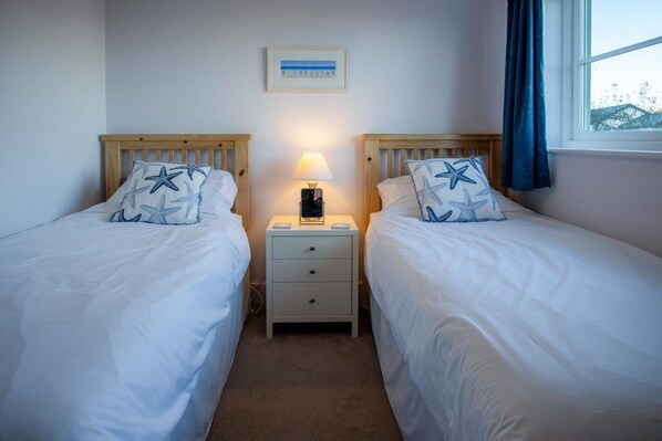 A twin room with two single beds, each with star-patterned pillows, a bedside table with a lamp, and a framed picture on the wall. - 13 Rhodewood House - 2 Bedroom Apartment - Saundersfoot (Saundersfoot)