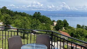 A balcony with wicker chairs and a glass table overlooking a lake, trees, and distant cityscape.