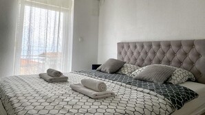 A neatly made bed with a patterned bedspread and pillows, a tufted headboard, and a window with sheer curtains.