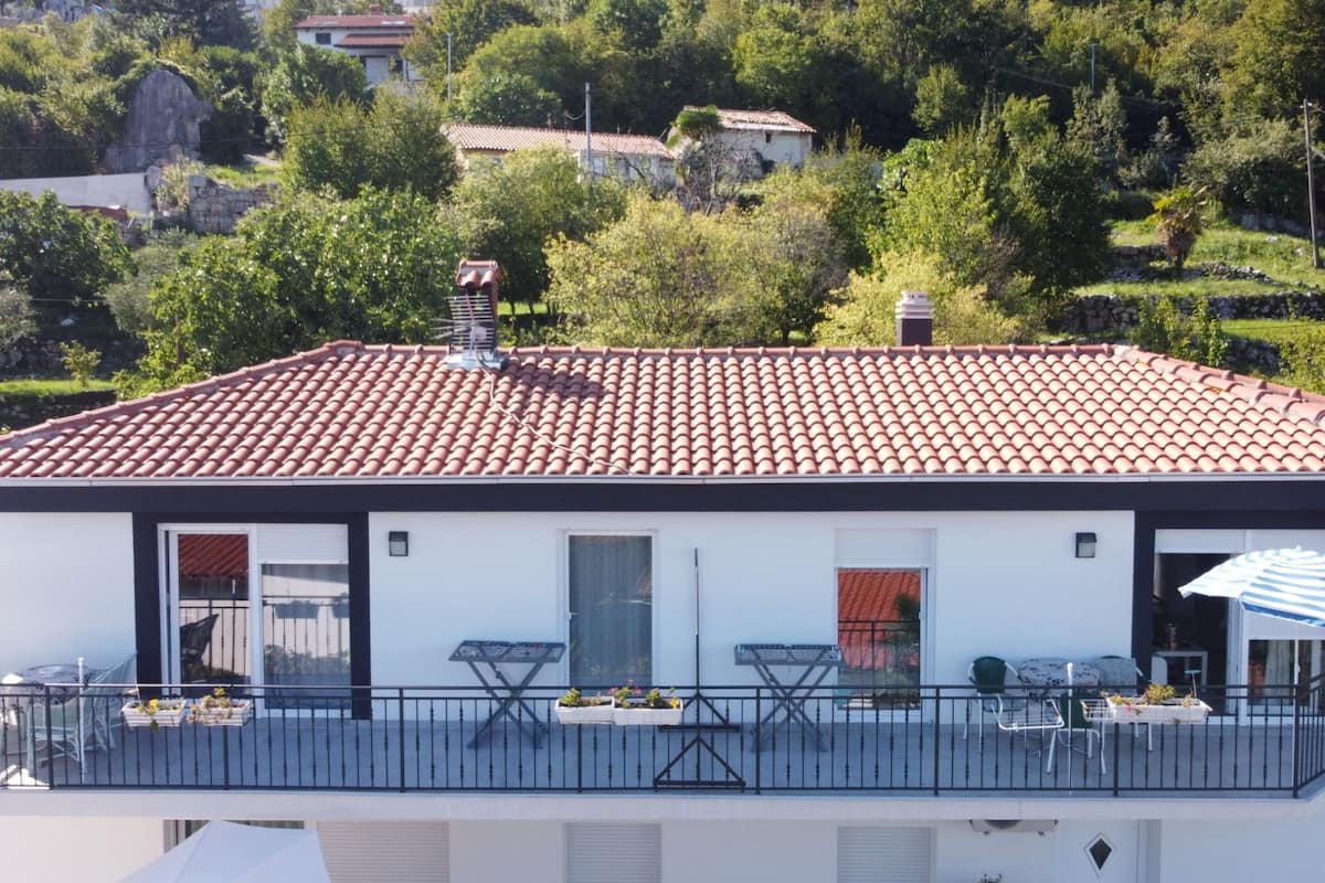 Un bâtiment moderne avec un toit recouvert de tuiles rouges, des murs blancs et un balcon agrémenté de mobilier d’extérieur.