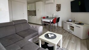 A compact living space with a grey sofa, a dining table with red chairs, a small white table with a bowl, a TV mounted on the wall, and a kitchenette in the background.