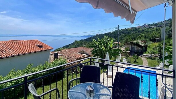 A balcony with a table and chairs, overlooking a swimming pool and a view of the sea and hills.