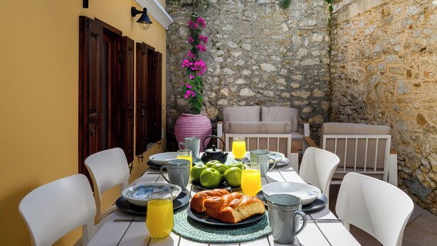 Une table à manger dressée avec des assiettes, un pain, des pommes et une carafe de jus, entourée de chaises blanches dans un espace extérieur aux murs de pierre.