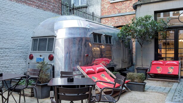Une caravane ancienne avec un lit rouge et une table entourée de chaises à l’avant.