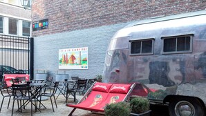 Une caravane ancienne est garée devant un bâtiment en briques, sur lequel on peut voir une enseigne indiquant la présence d’un bar à vin et d’un hôtel. Des espaces de détente extérieurs, avec des chaises et des tables, sont aménagés.