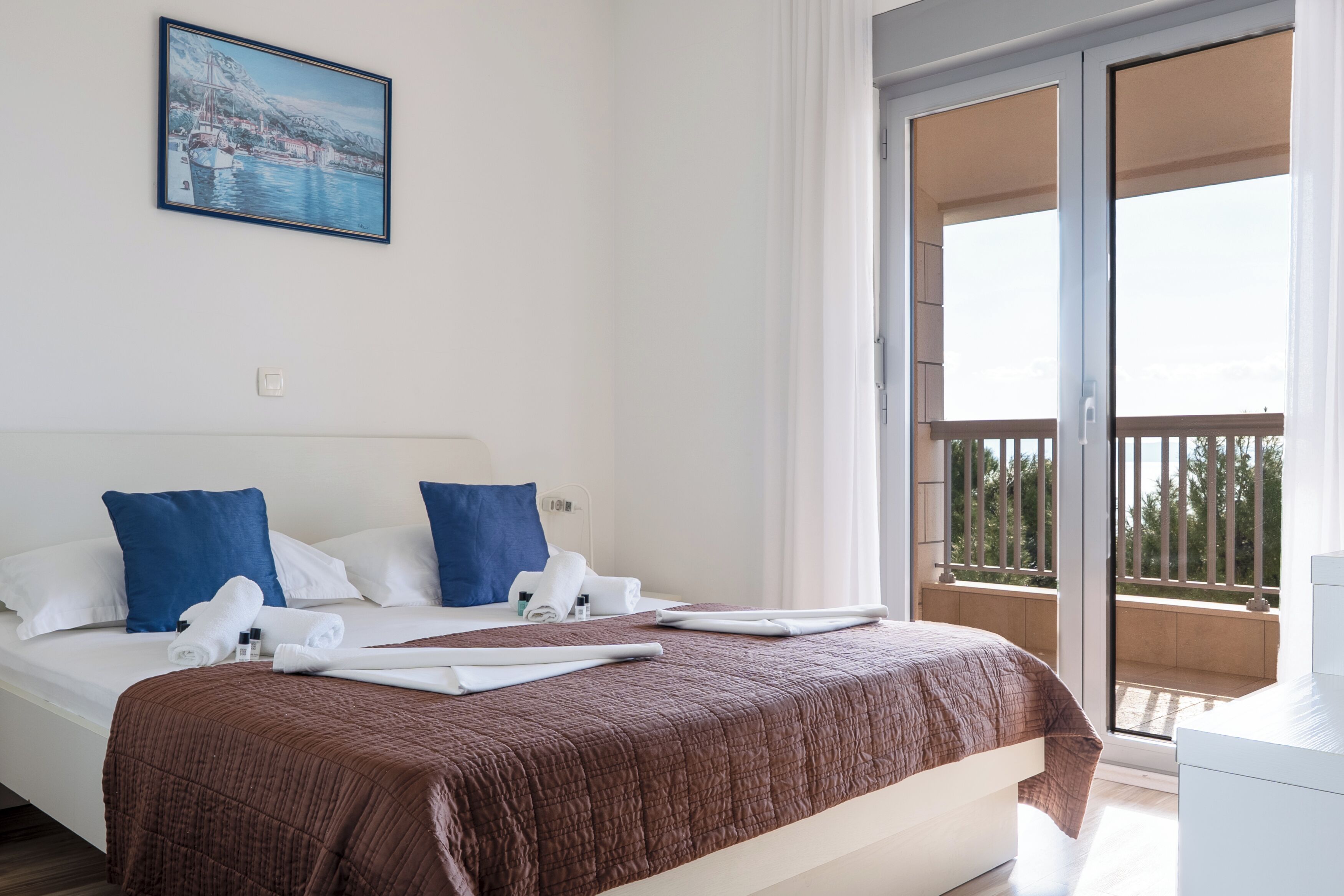 A hotel room with a bed, blue pillows, a brown bedspread, and a balcony with a view.