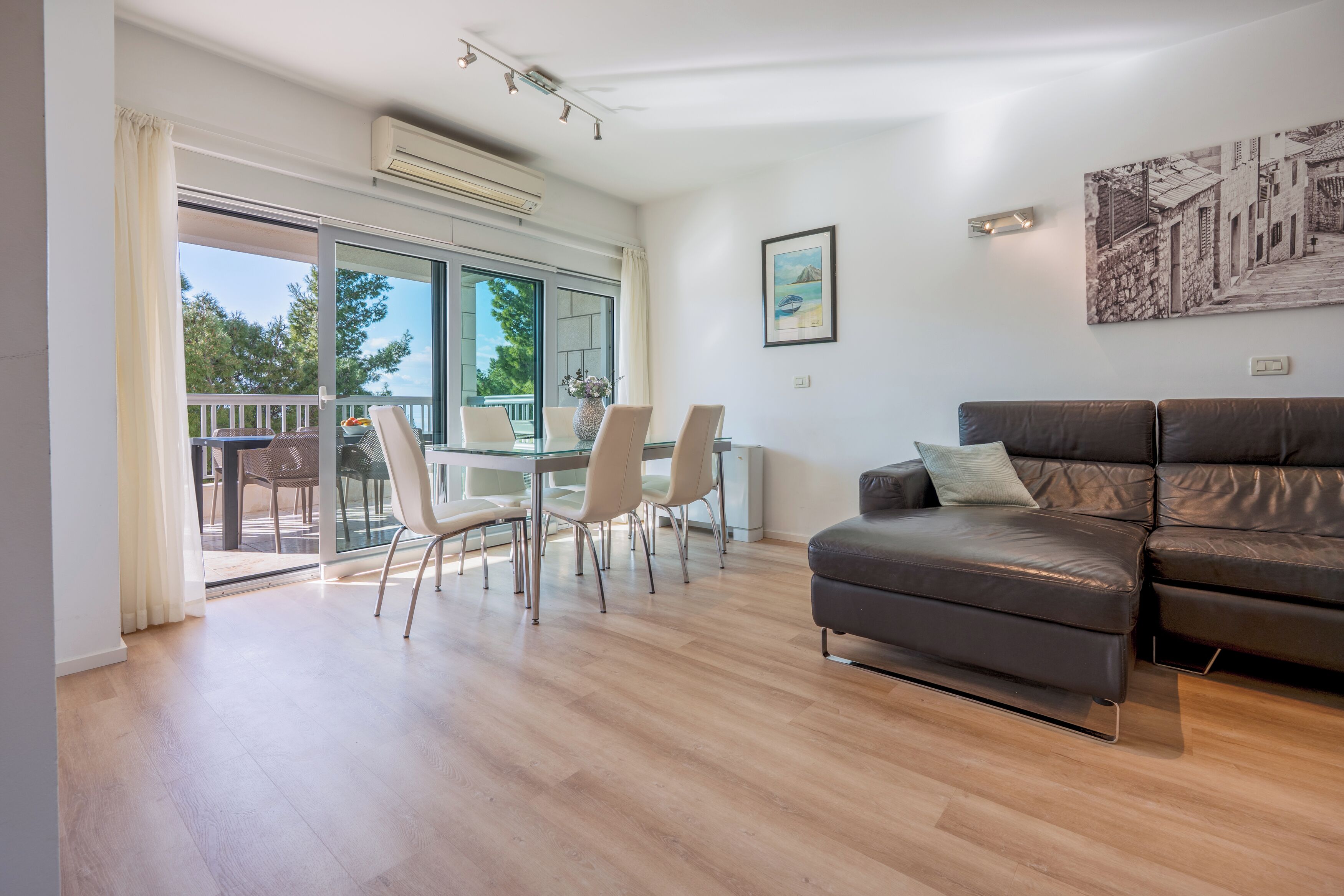 A modern living room with a leather sofa, a glass dining table with chairs, and a balcony with a view of trees and a building.